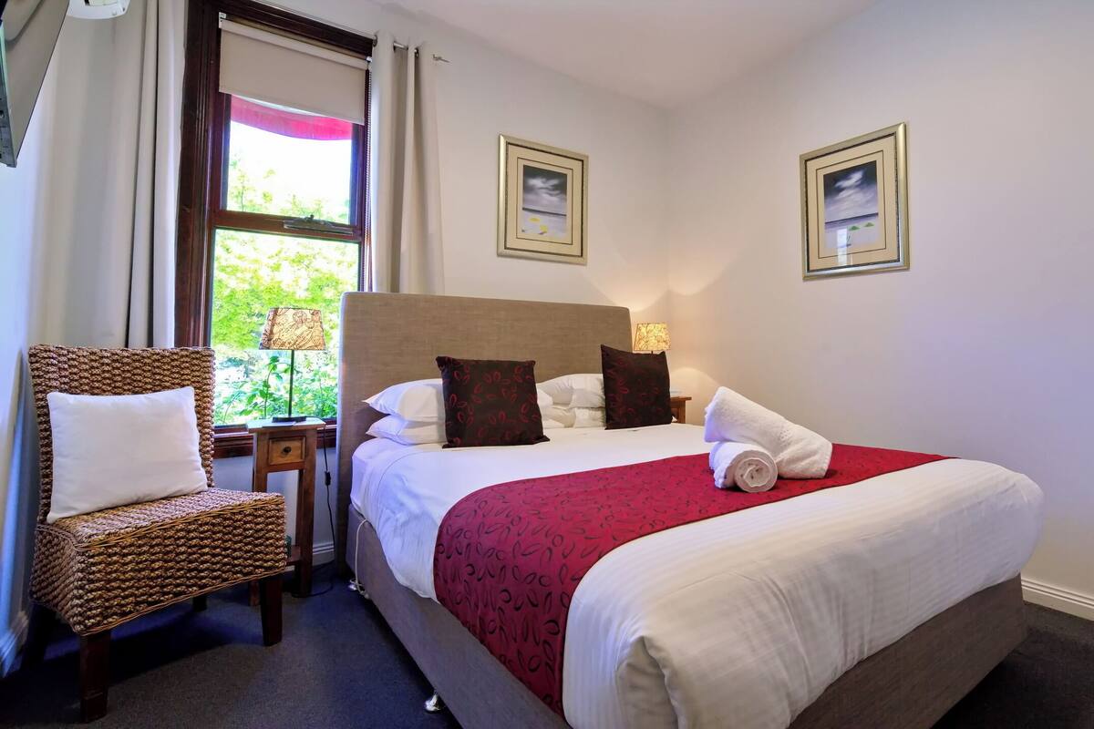 A neatly made bed with white and red linens, a wooden headboard, and two pillows. There is a wicker chair with a white cushion, a small table with a lamp, and two framed pictures on the wall.