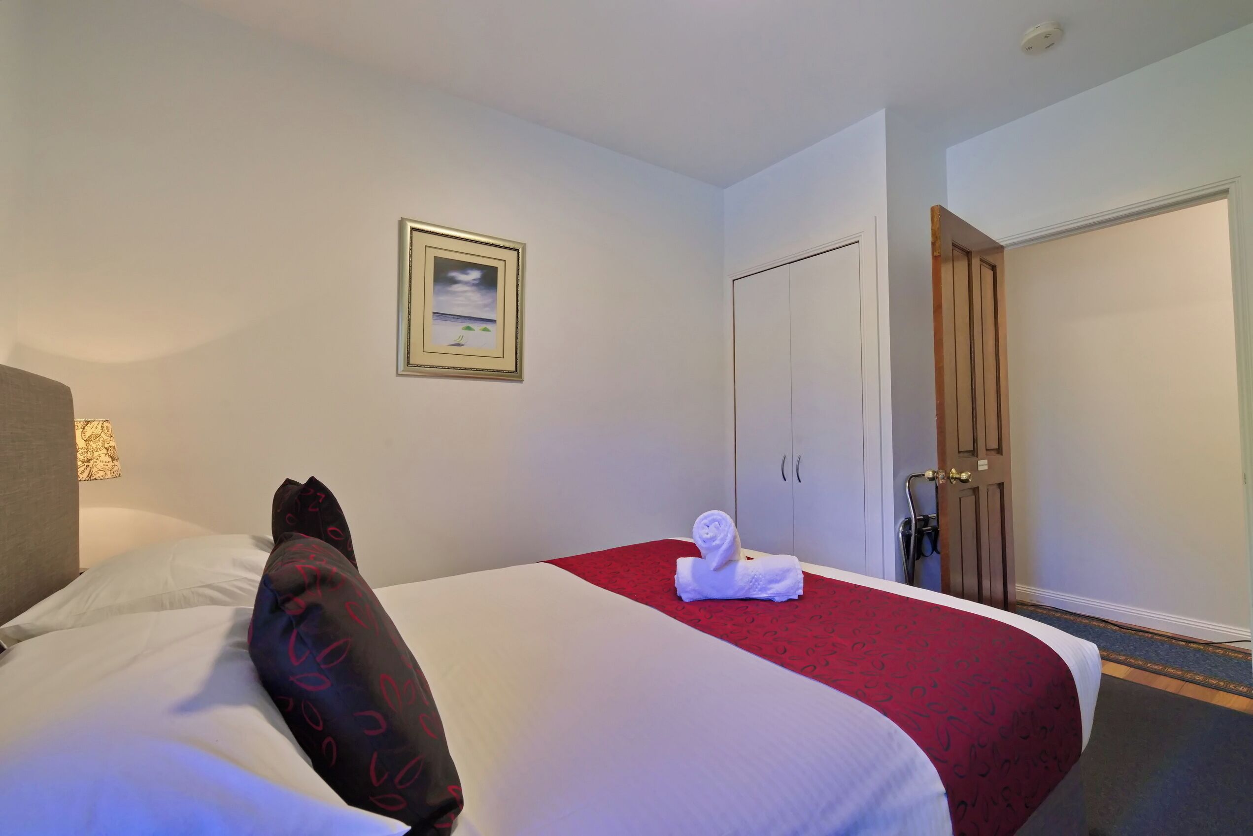 A hotel room with a bed, a red bedspread, a white pillow, a framed picture on the wall, and a door with a towel on it.