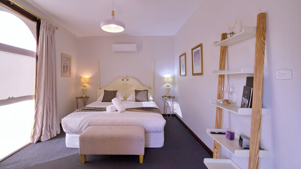 Una habitación de hotel con una cama grande, lámparas de noche, un estante de madera con libros y vasos, y una ventana con cortinas.