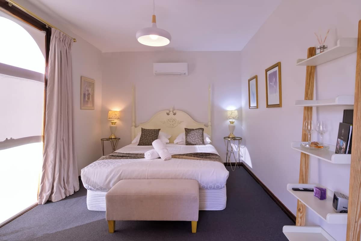A hotel room with a large bed, bedside lamps, a wooden shelf with books and glasses, and a window with curtains.