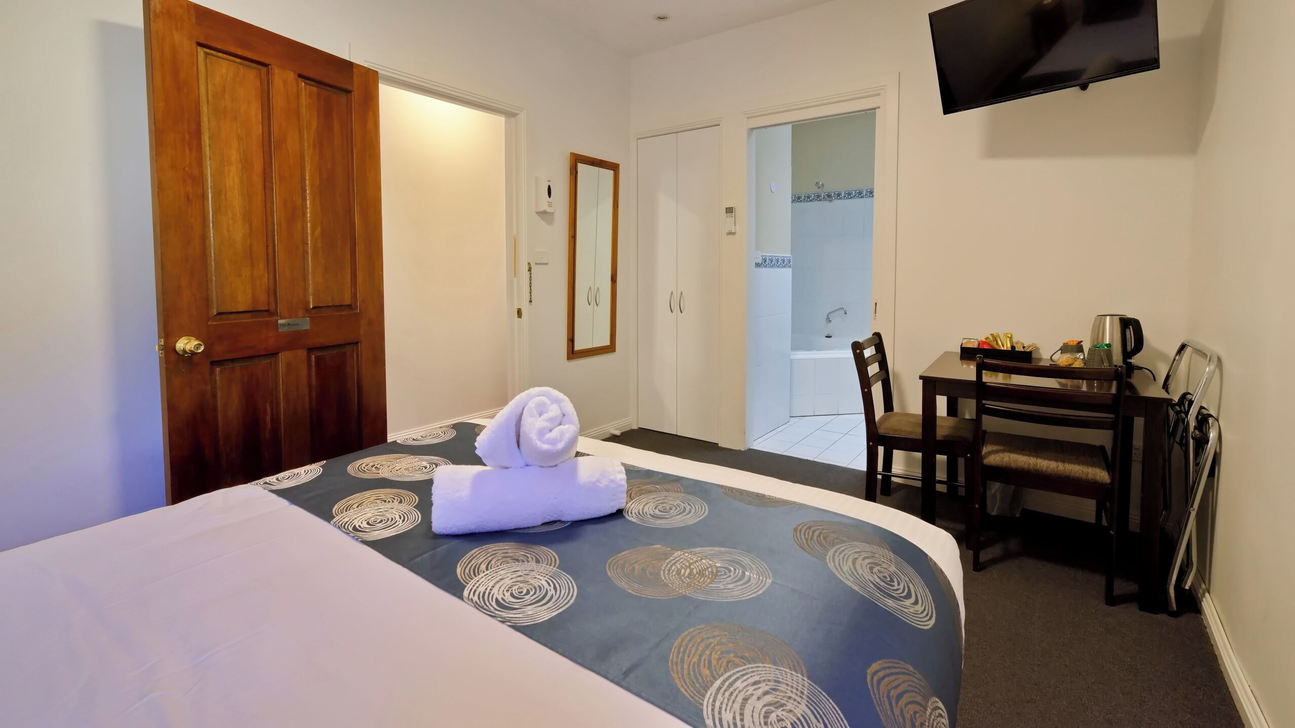 A hotel room with a bed, a desk, a chair, a TV, and a bathroom door.
