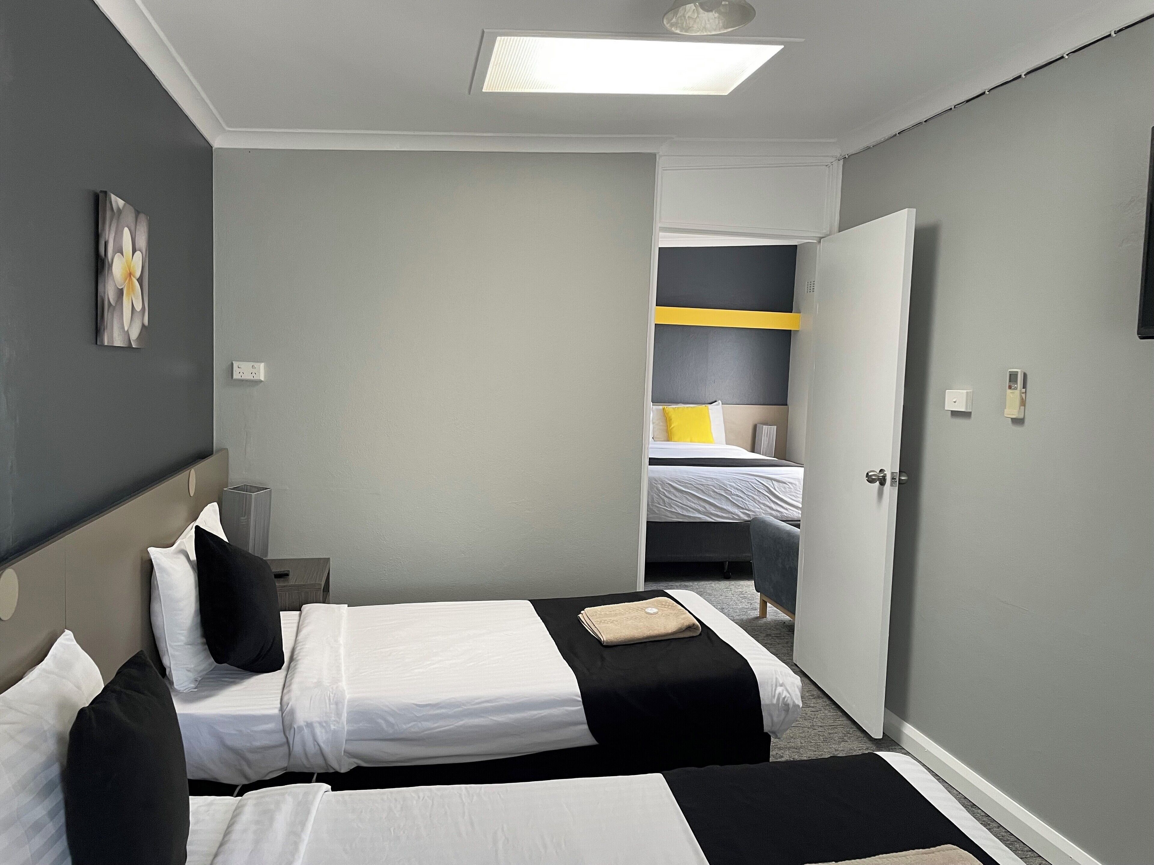 A hotel room with two beds, a bedside table, a lamp, a painting, and a door leading to another room.