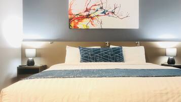 Quarto de hotel com uma cama grande, duas luminárias de cabeceira, uma pintura abstrata na parede e uma cabeceira com torneira.