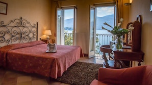 A bedroom with a bed, a nightstand, a lamp, a chair, a small table, a vase with flowers, and a view of a lake and mountains through a balcony door. - Hotel Aquadolce (Verbania)