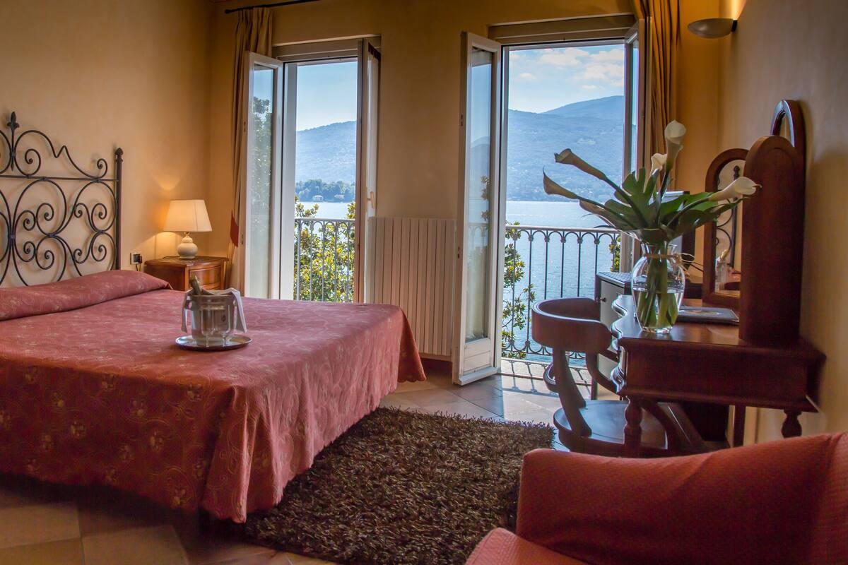 A bedroom with a bed, a nightstand, a lamp, a chair, a small table, a vase with flowers, and a view of a lake and mountains through a balcony door.