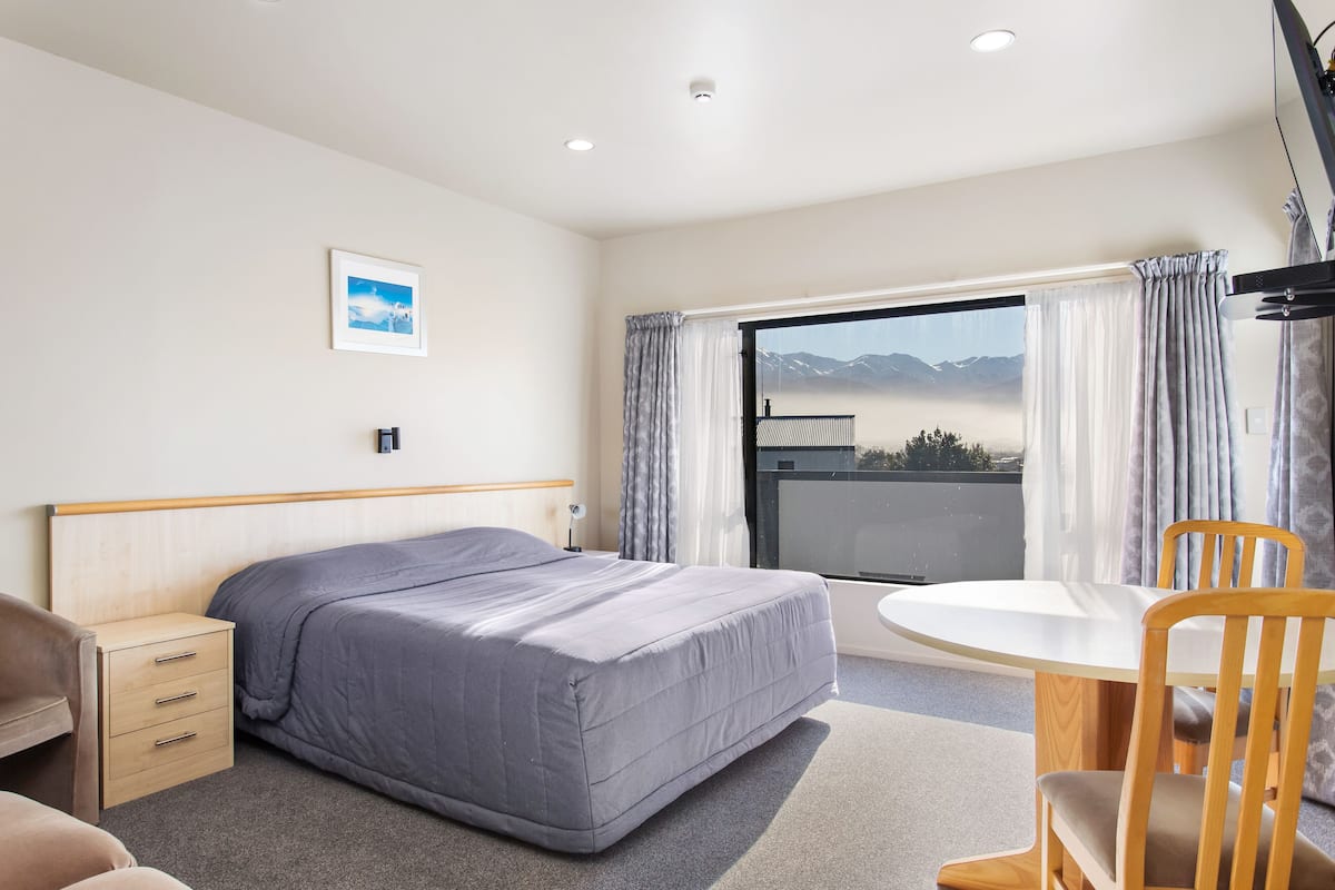 A hotel room with a bed, bedside table, chair, and a view of mountains through the window.