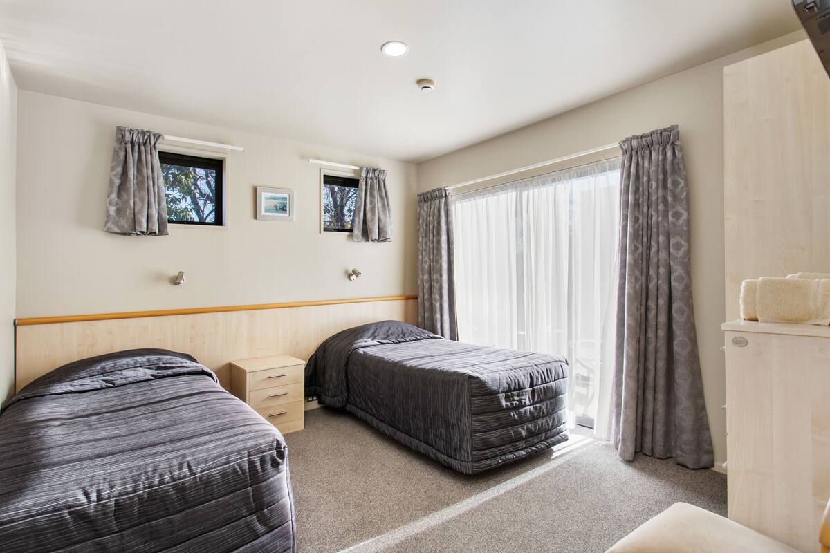A hotel room with two single beds, a wooden headboard, a small bedside table, a wardrobe, and a large window with curtains.