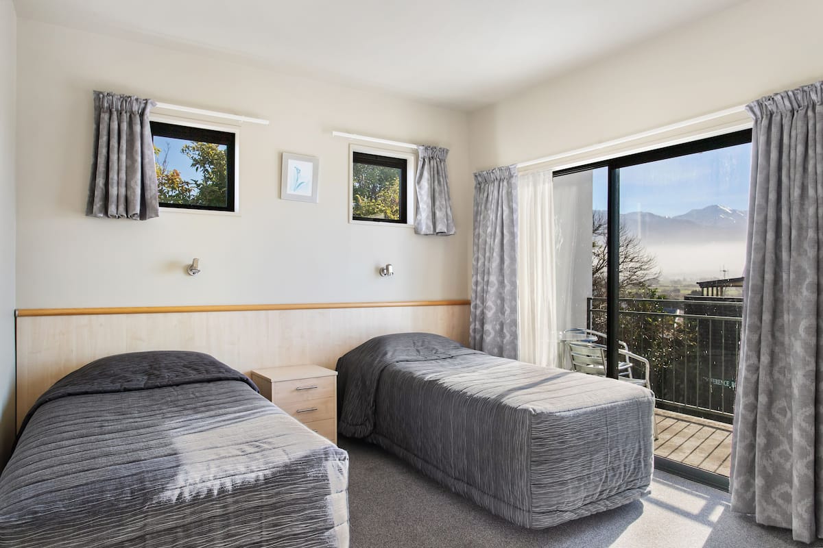 A hotel room with two beds, a small desk, and a balcony with a view of mountains.