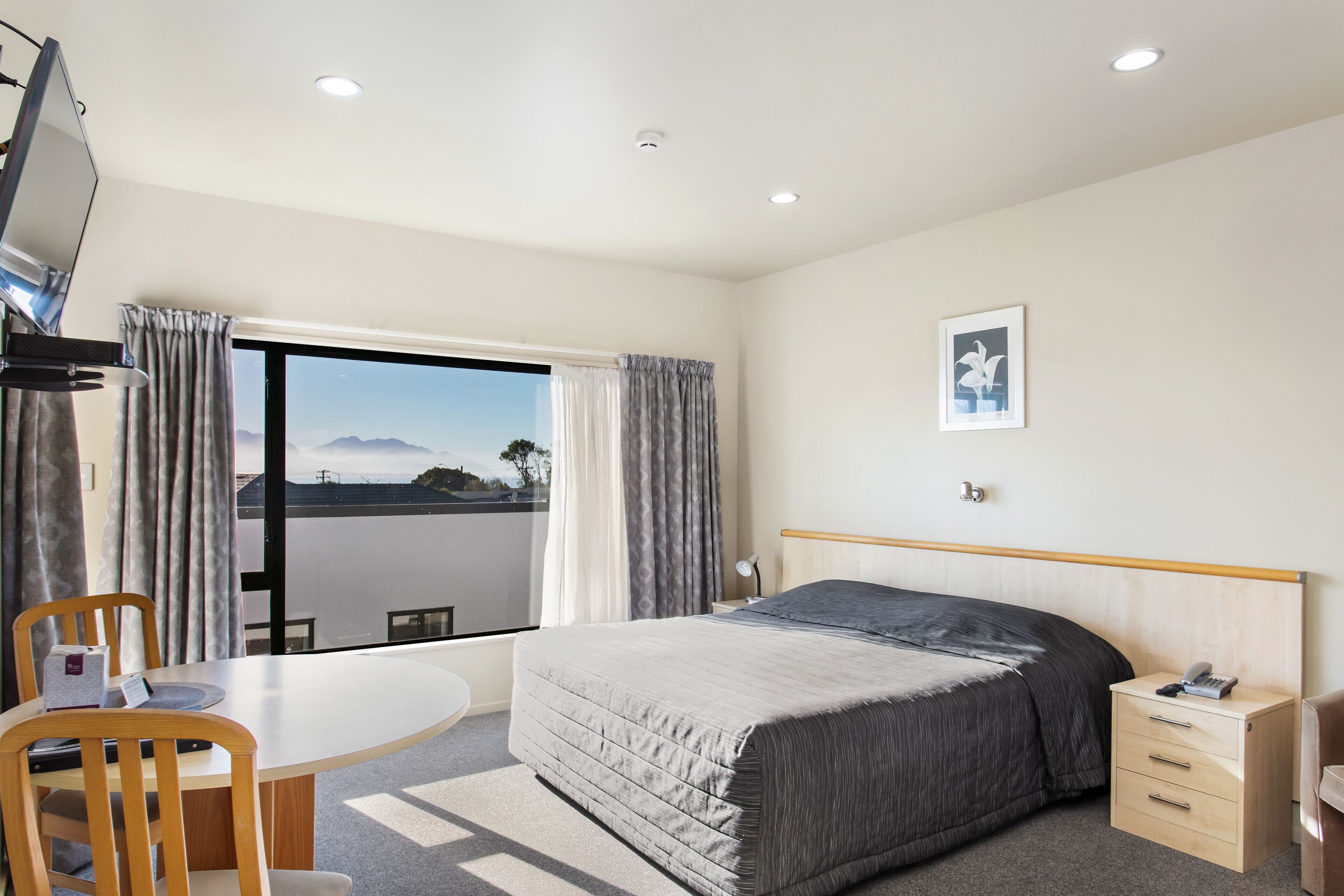 Une chambre d’hôtel comprenant un lit, un bureau, une chaise, une télévision, une table de chevet, une lampe et une fenêtre avec des rideaux.