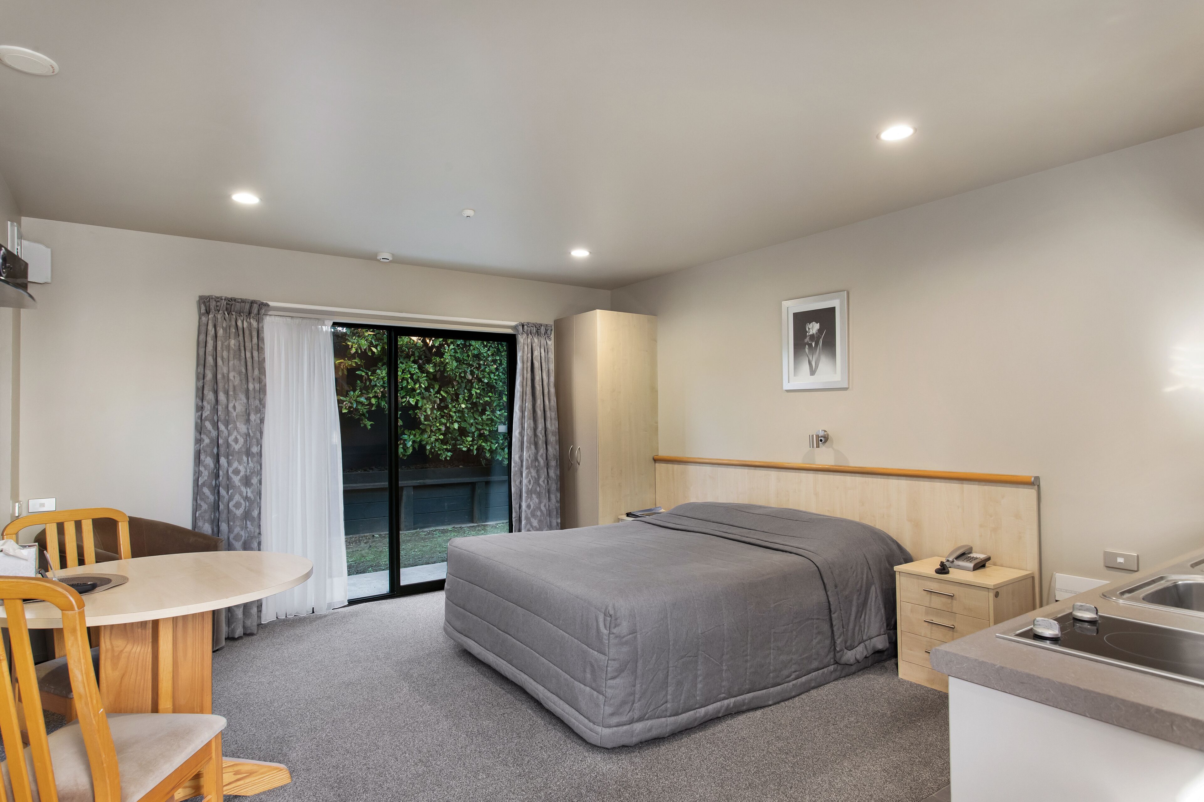 A modern hotel room with a bed, a small desk, a chair, a window with curtains, a sink, and a framed picture on the wall.