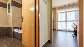 Baño de hotel con ducha, lavabo e inodoro. Una puerta de madera da a una habitación con cama y ventana.
