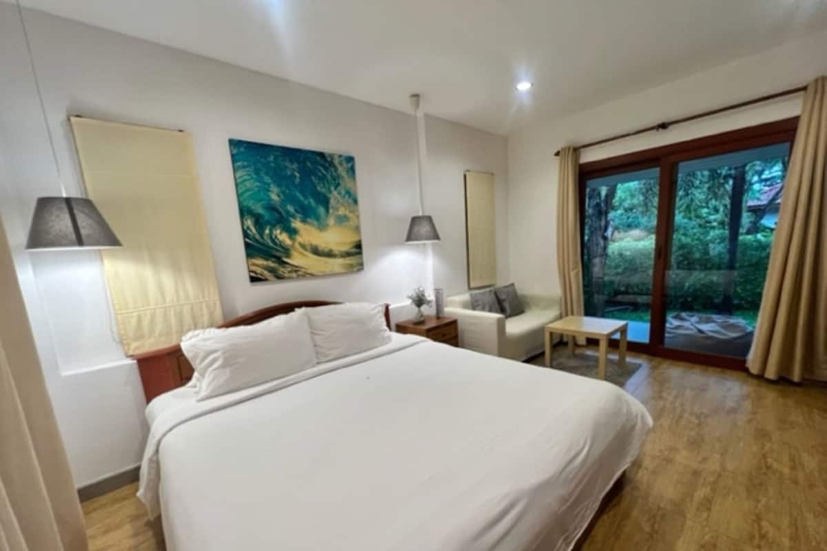 Ett hotellrum med en säng, en soffa, ett bord och ett stort fönster med utsikt över grönområden.