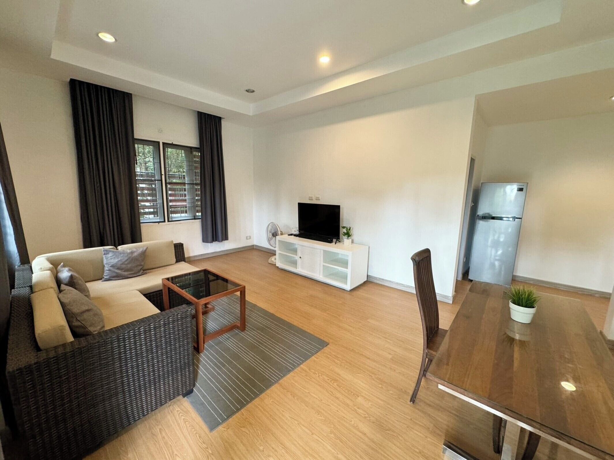 A modern living room with a sofa, a coffee table, a TV on a white cabinet, a dining table with chairs, and a refrigerator.