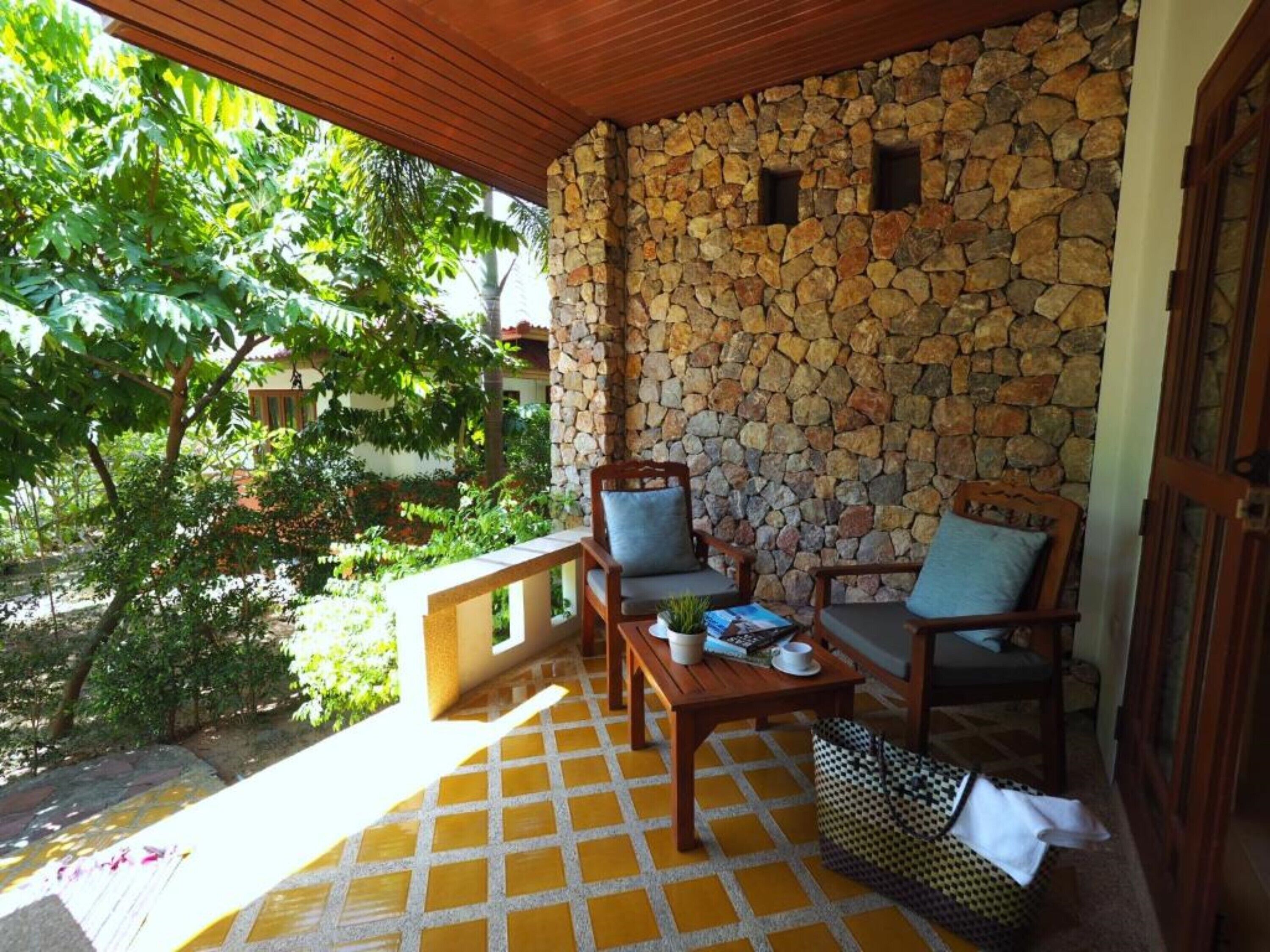 A covered outdoor seating area with stone walls, wooden furniture, and a view of greenery.