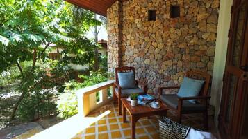 A covered outdoor seating area with stone walls, wooden furniture, and a view of greenery.