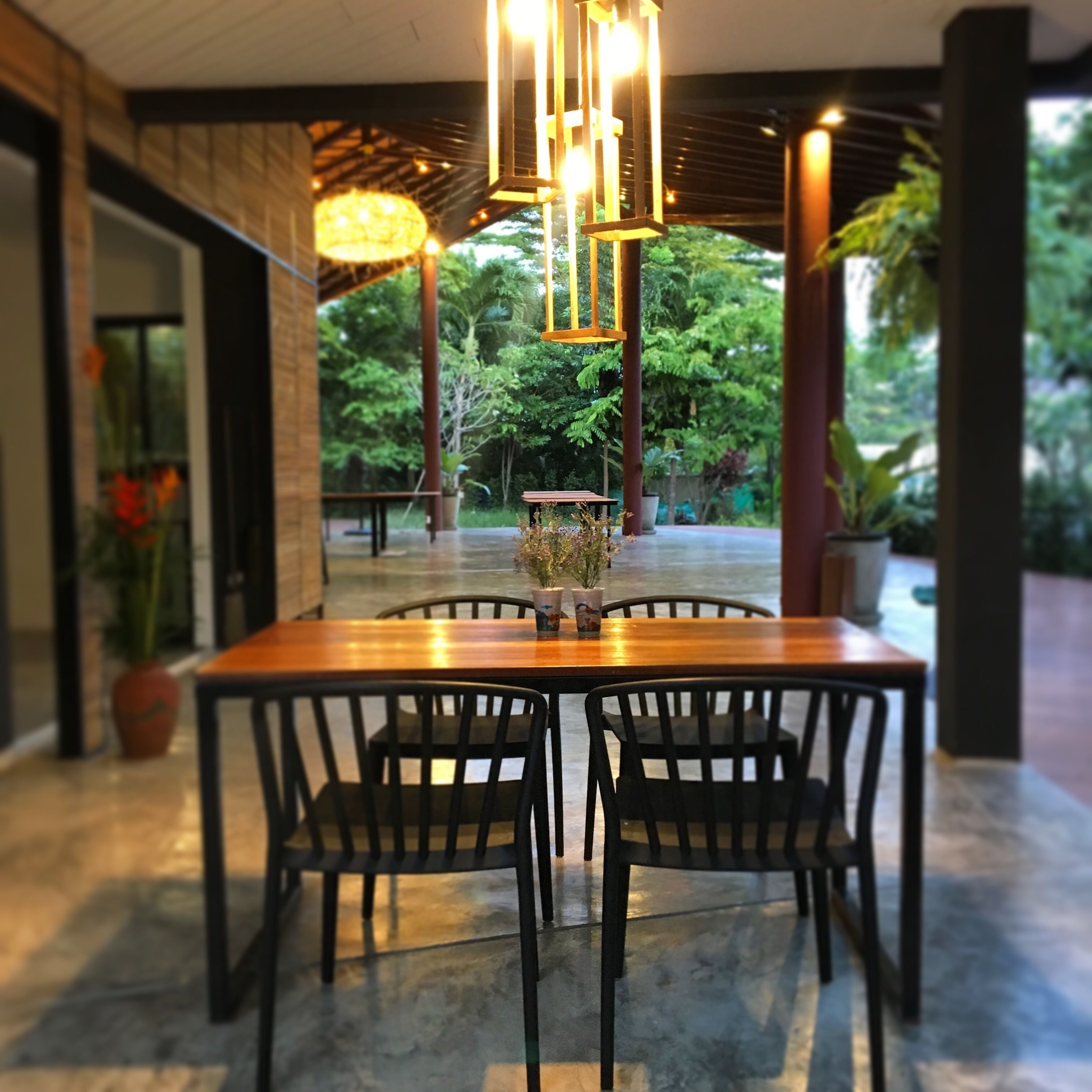 A dining area with a wooden table and black chairs, surrounded by potted plants and a view of greenery.
