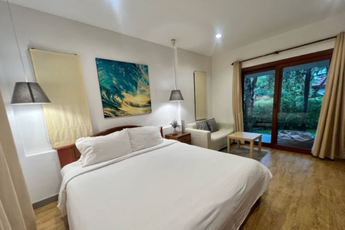 Ett hotellrum med en stor säng, ett litet bord, en stol och utsikt över grönområden genom en skjutdörr.