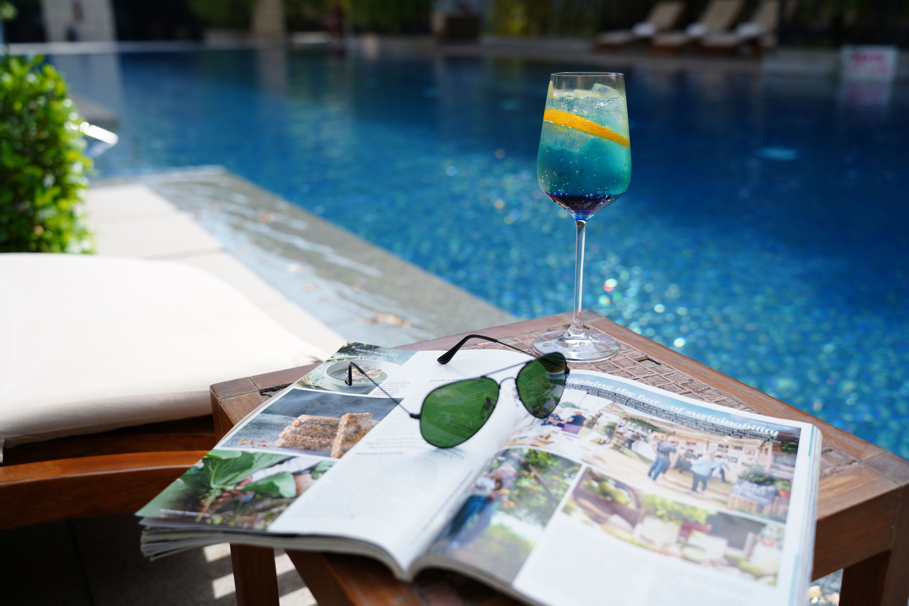 A poolside table with a drink, sunglasses, and a magazine.