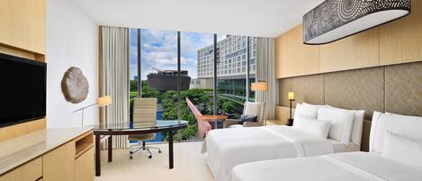 A hotel room with a large TV, a glass table, two beds, a chair, and a view of a building and greenery outside.