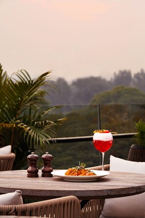 Une table sur laquelle se trouve une assiette de nourriture et un verre d’une boisson rouge, entourée de chaises et de coussins en osier.