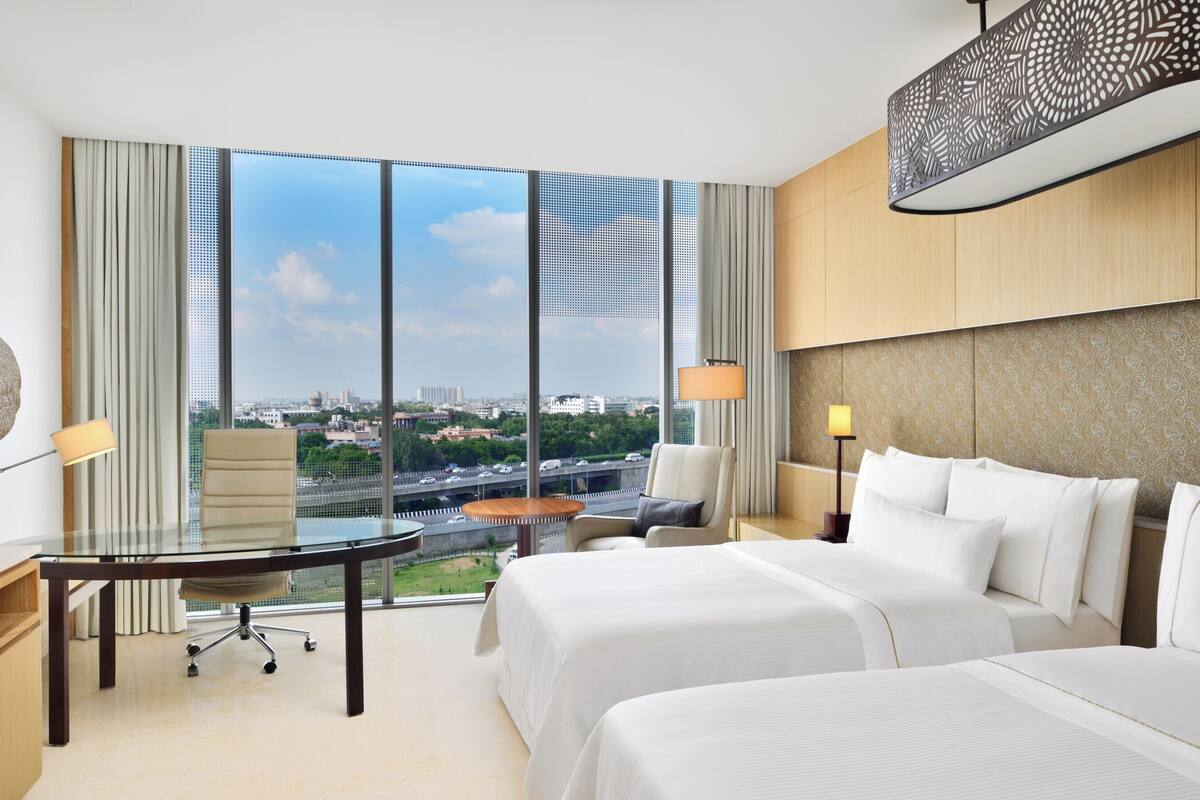 Quarto de hotel com uma grande janela, TV de tela plana, escrivaninha, duas camas e um objeto decorativo na parede.
