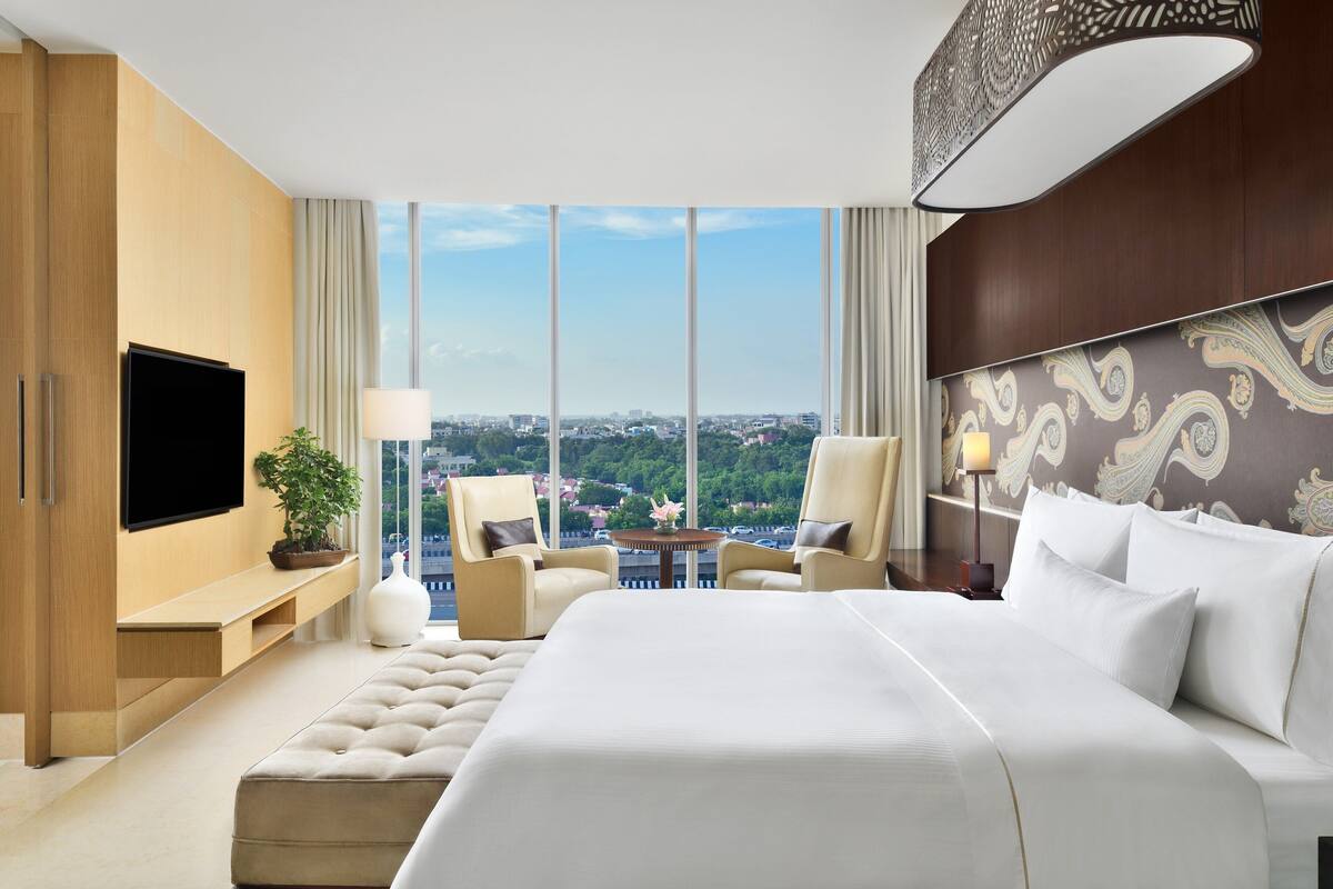 Quarto de hotel moderno com uma cama grande, duas poltronas, uma televisão e vista para a cidade.
