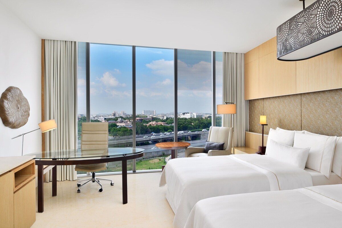 A hotel room with a large window, a desk, a chair, a bed, and a nightstand with lamps.