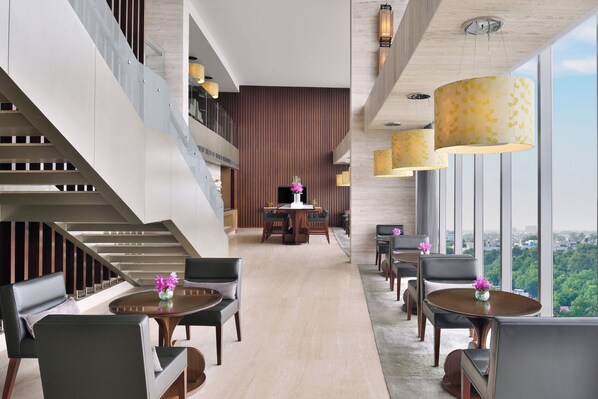 Un moderno vestíbulo de hotel con una escalera, zona de comedor y amplias ventanas con vista a la ciudad.