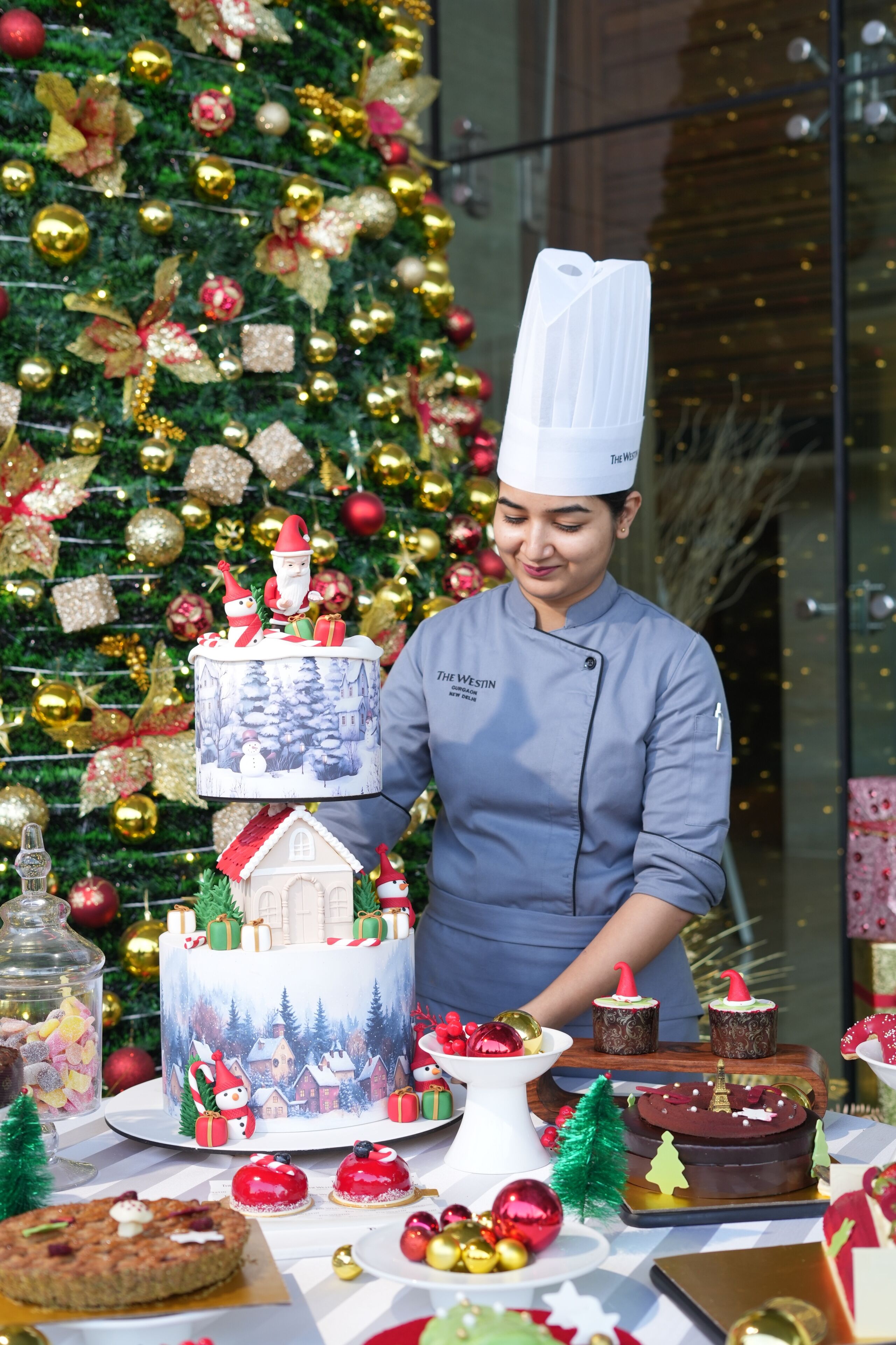 A chef in a uniform with a name tag, standing in front of a decorated Christmas tree and a multi-tiered cake with festive decorations.