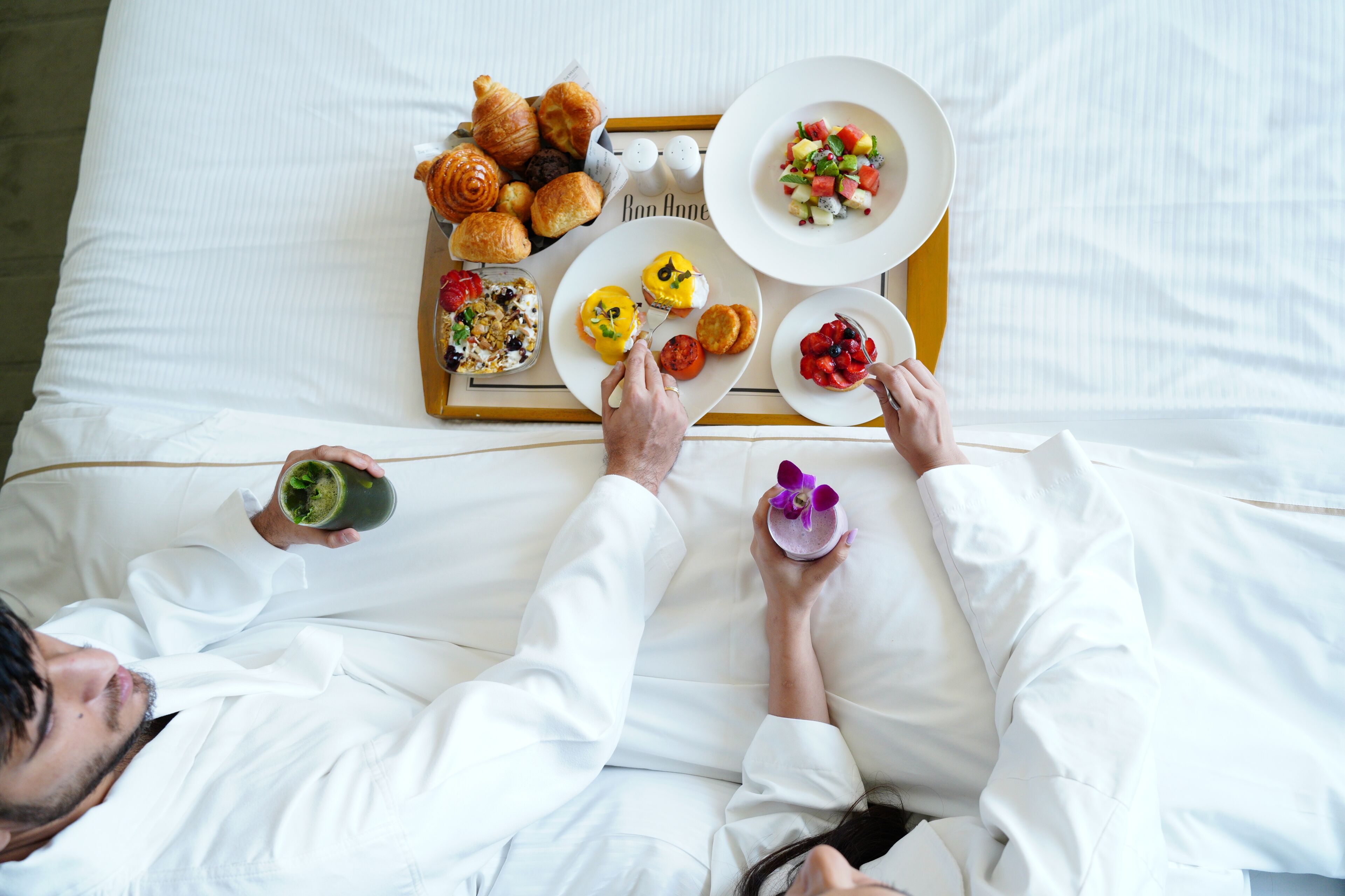 Una cama con una bandeja de desayuno que incluye cruasanes, una ensalada de frutas y una bebida verde.
