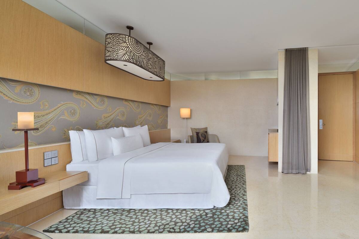Quarto de hotel moderno com uma cama grande, uma mesa de cabeceira, um abajur e um tapete estampado.