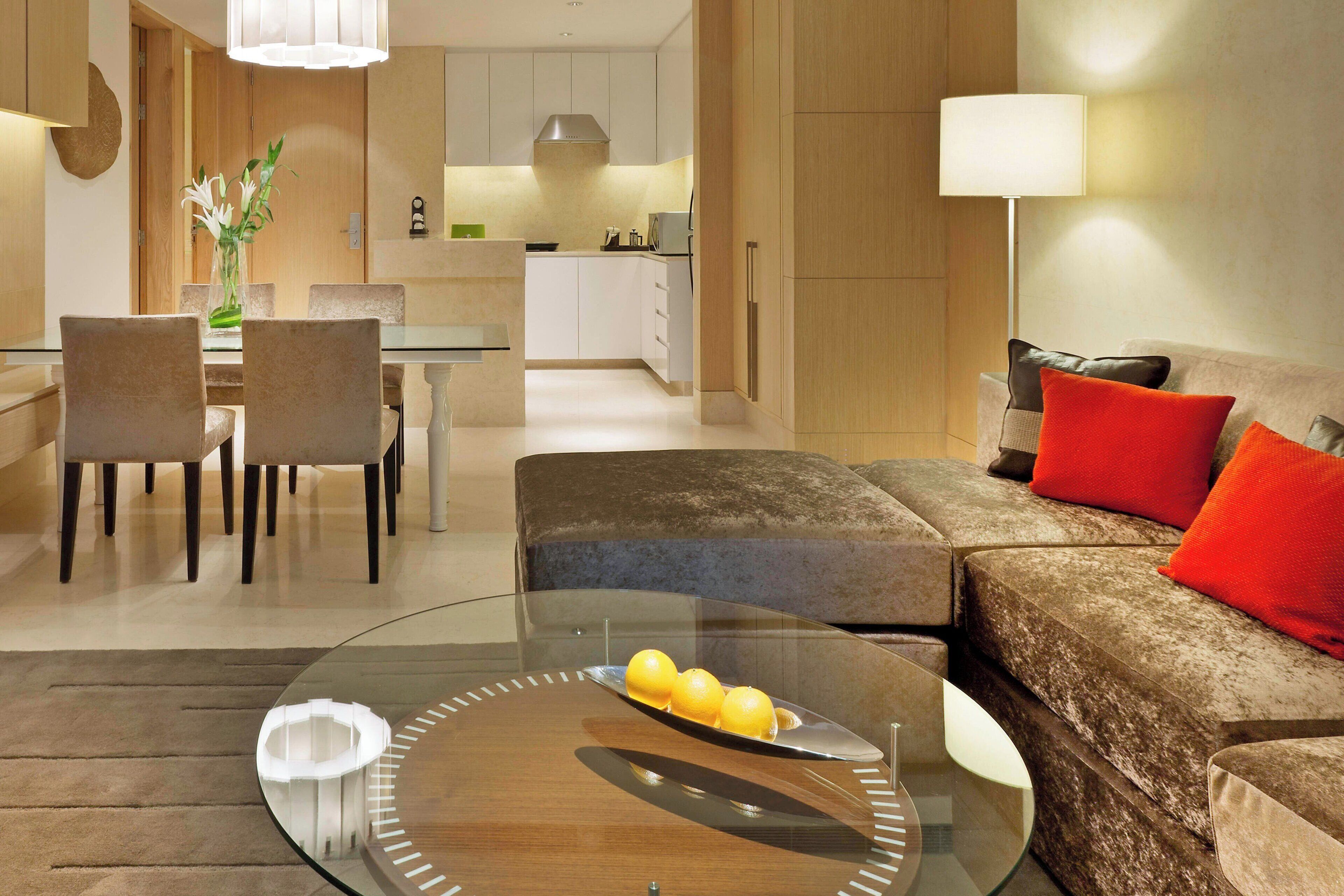 A modern living room with a glass coffee table, a sofa with red cushions, and a dining area with chairs and a vase of flowers.