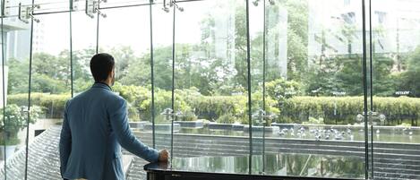 A man in a blue suit walking towards a grand piano in a spacious, modern room with large windows overlooking a garden.
