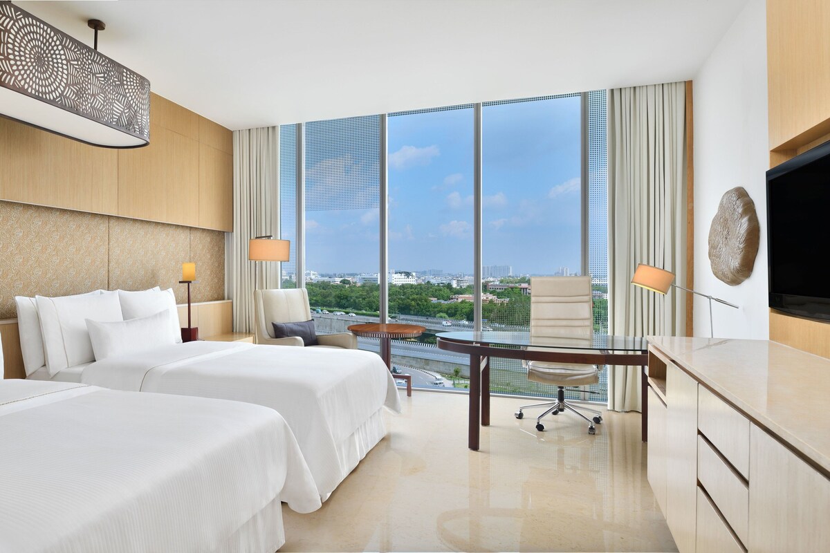 A hotel room with two beds, a desk, a chair, and a large window offering a city view.