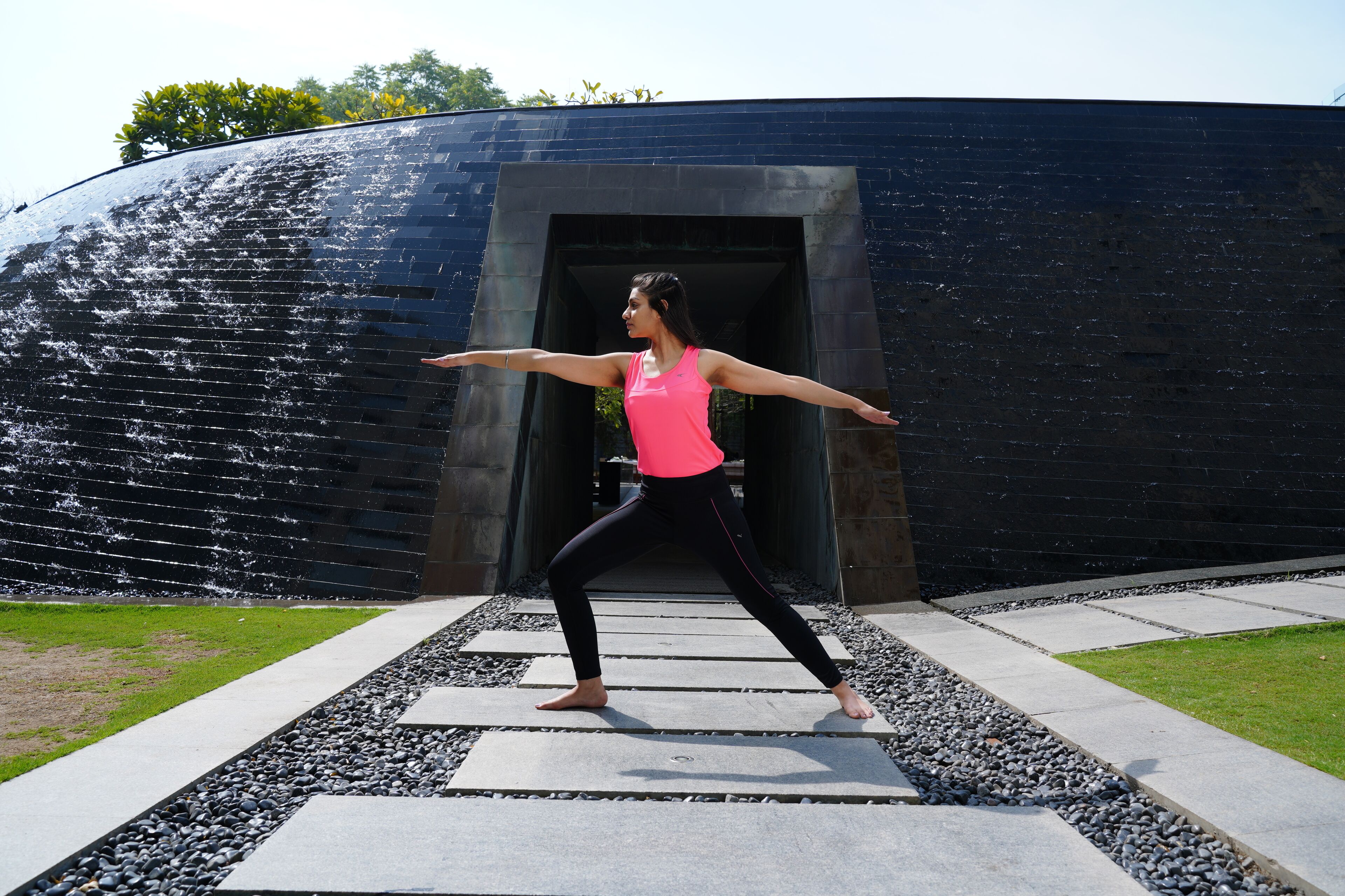 Une personne effectue une posture de yoga sur un chemin de béton, avec un mur en briques noires et de la verdure en arrière-plan.