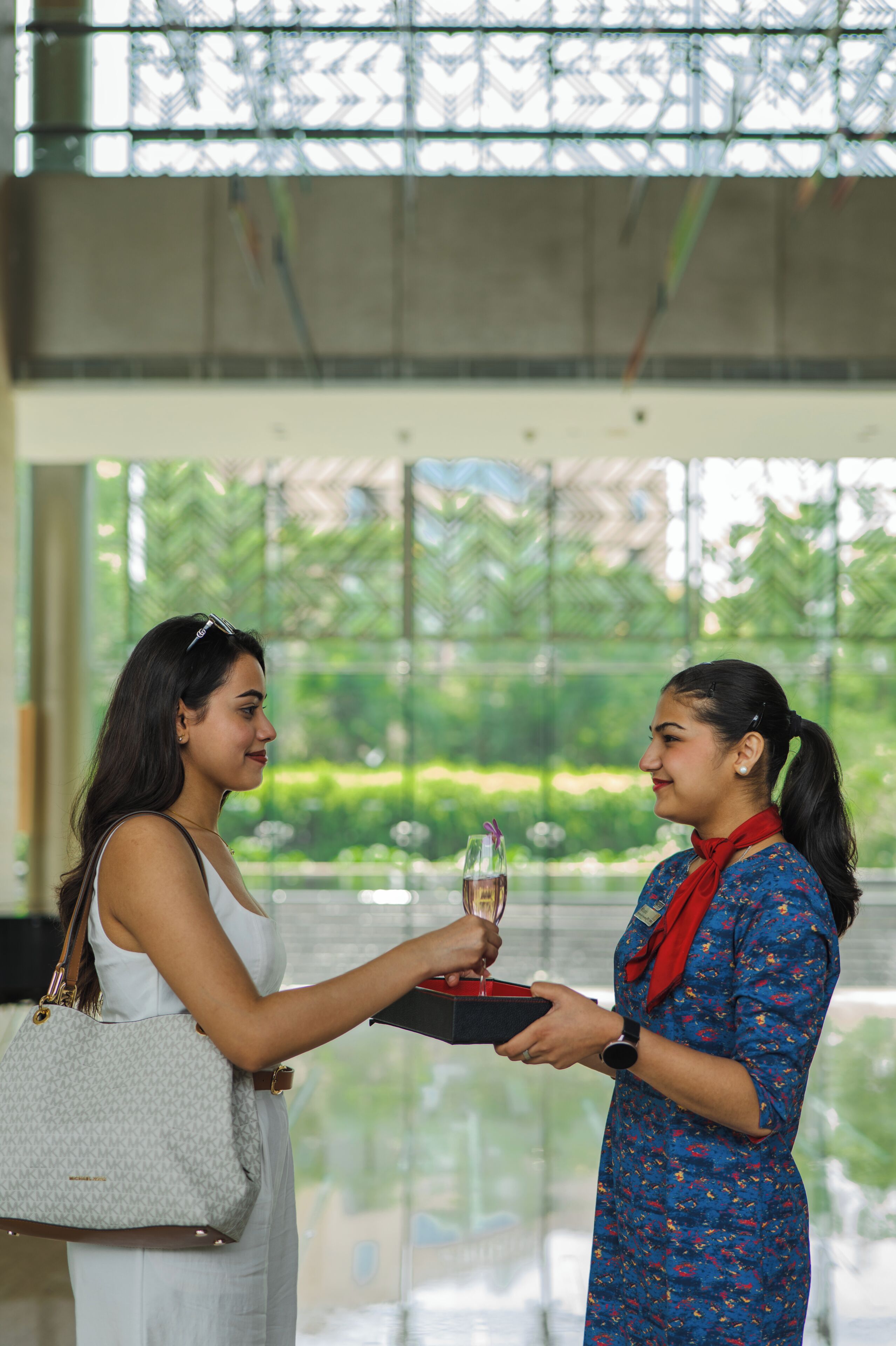 Dos mujeres en un espacio interior amplio y luminoso, con grandes ventanales; una sostiene una bebida y la otra, una bandeja.