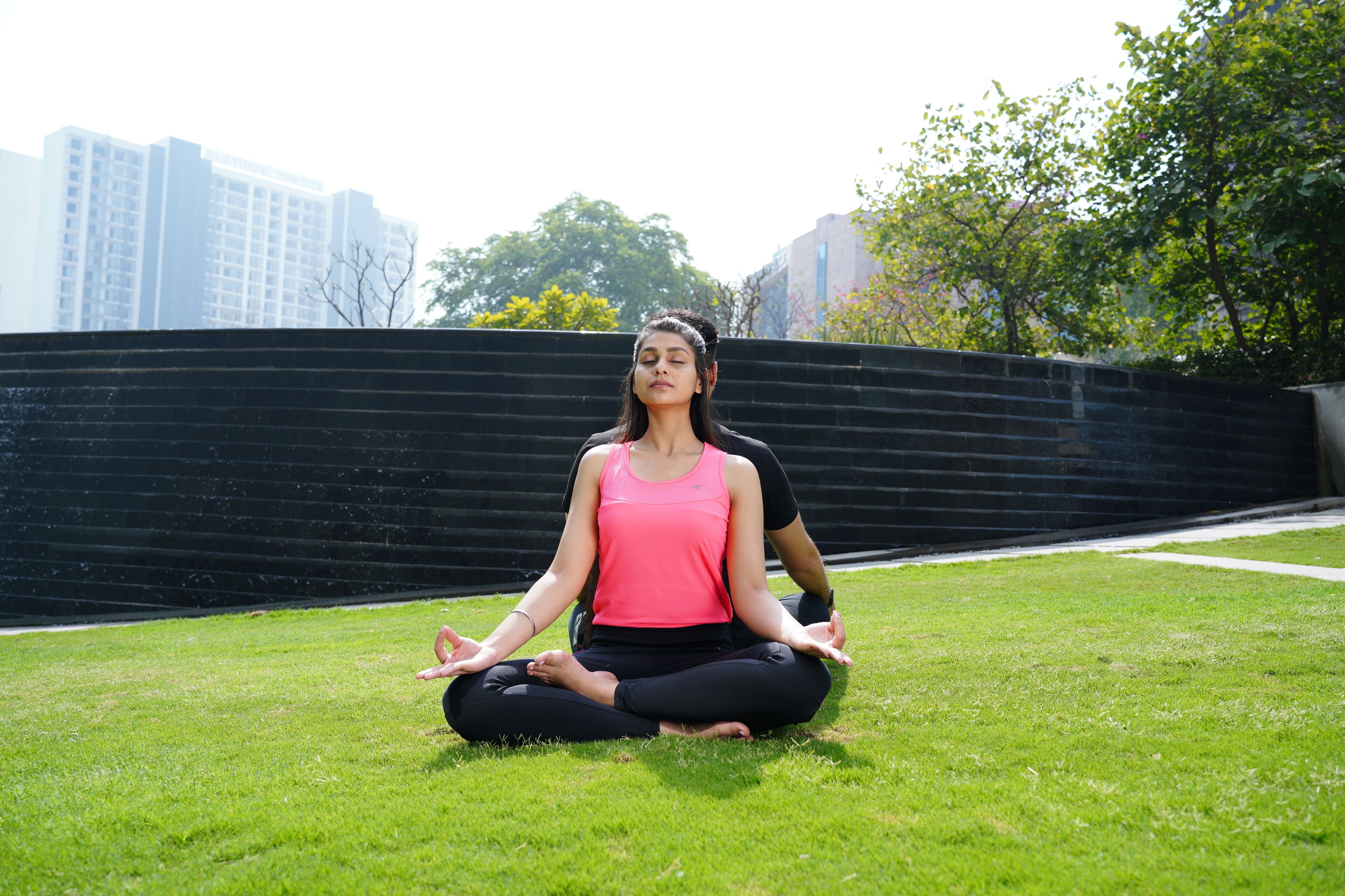 A person sitting cross-legged on grass in an outdoor area with a black wall and buildings in the background.