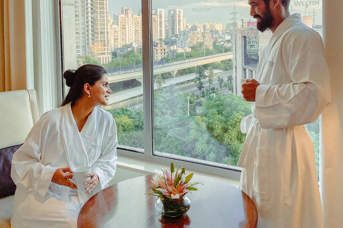 Quarto de hotel com uma janela ampla, uma mesa redonda com um vaso de flores e duas pessoas vestindo roupões brancos.