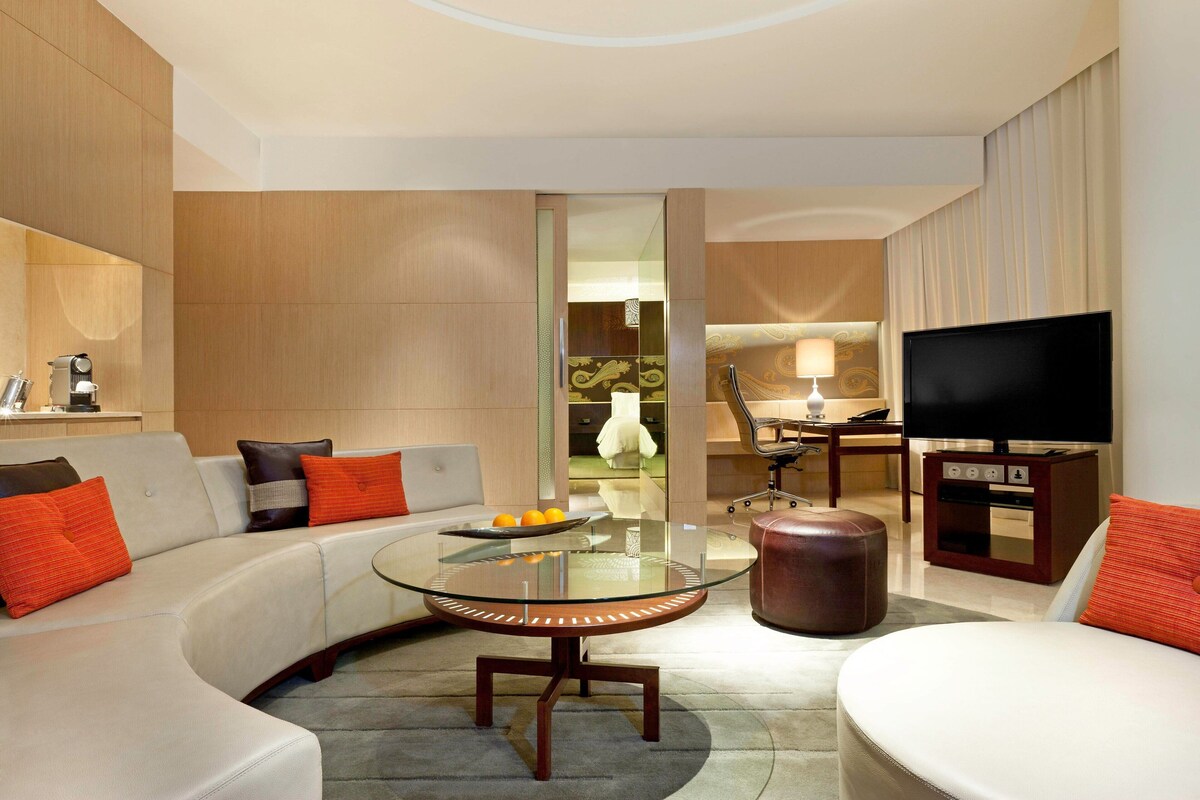 A modern living room with a glass-top coffee table, a sofa, a TV, and a kitchen area in the background.
