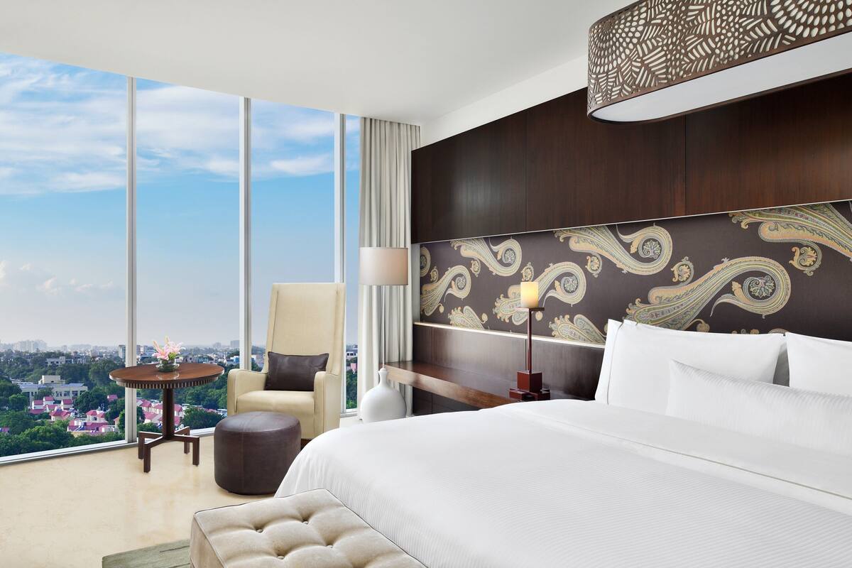 Quarto de hotel moderno com uma cama grande, uma mesa redonda, uma cadeira e vista para o horizonte urbano através de amplas janelas.