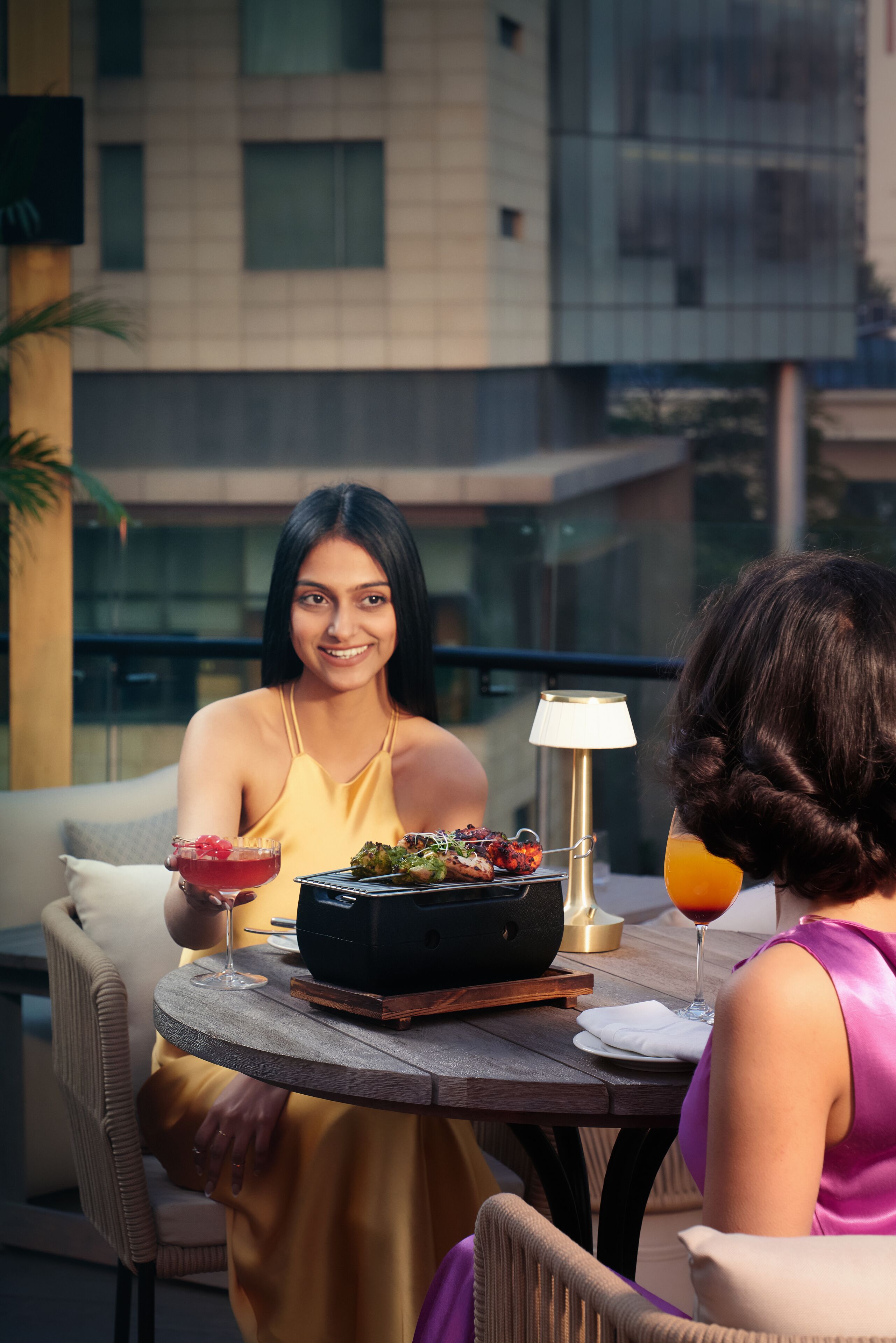 A dining area with a table set for two, featuring a bowl of food and drinks, a lamp, and a modern building in the background.