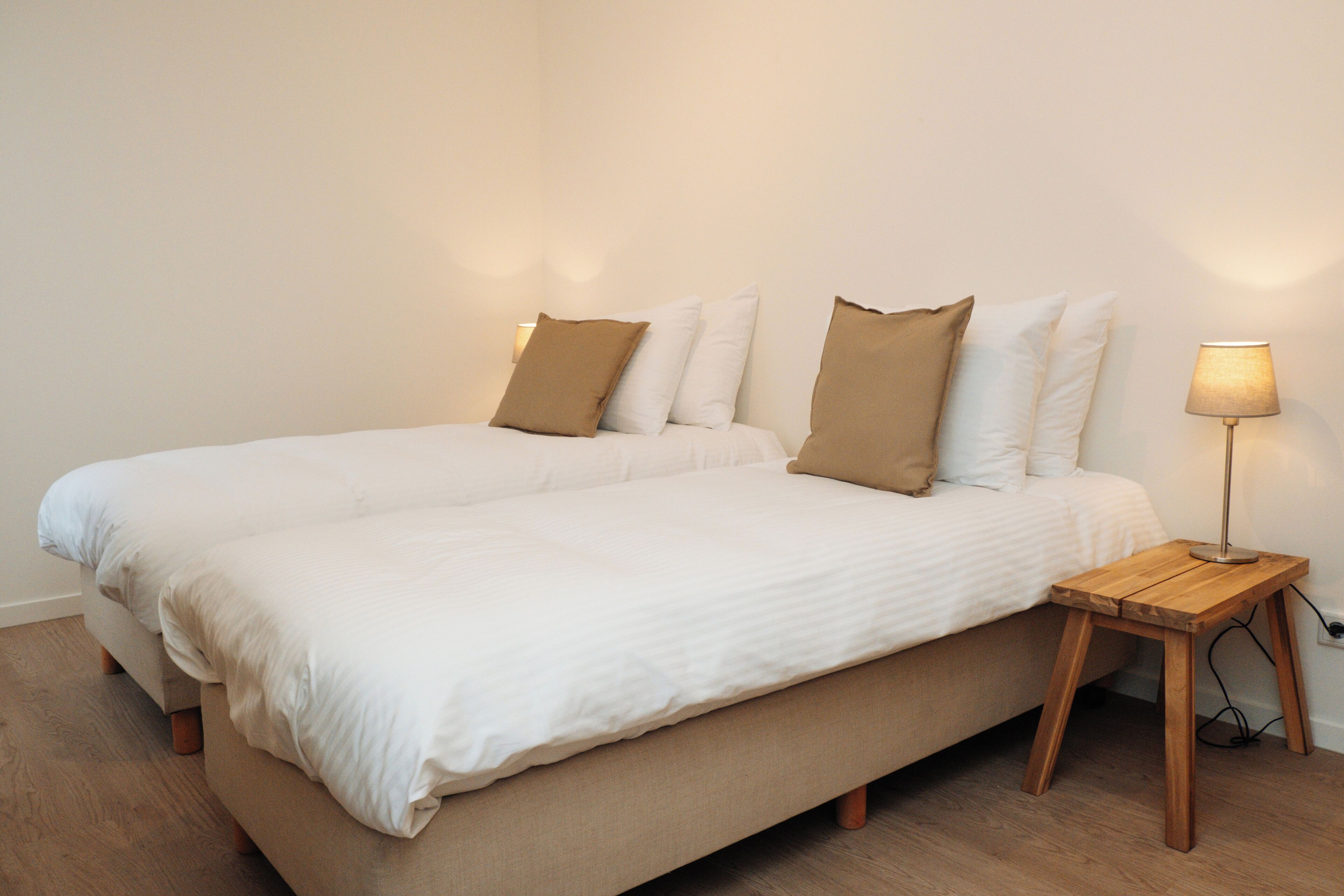 Een tweepersoonsbed met wit en bruin beddengoed, een houten nachtkastje met een lamp, en een houten vloer.