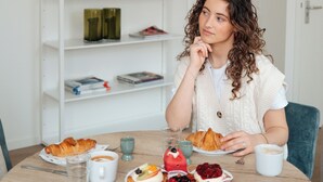 Una persona sentada a una mesa redonda con comida y bebidas, una estantería al fondo y una puerta a la derecha.