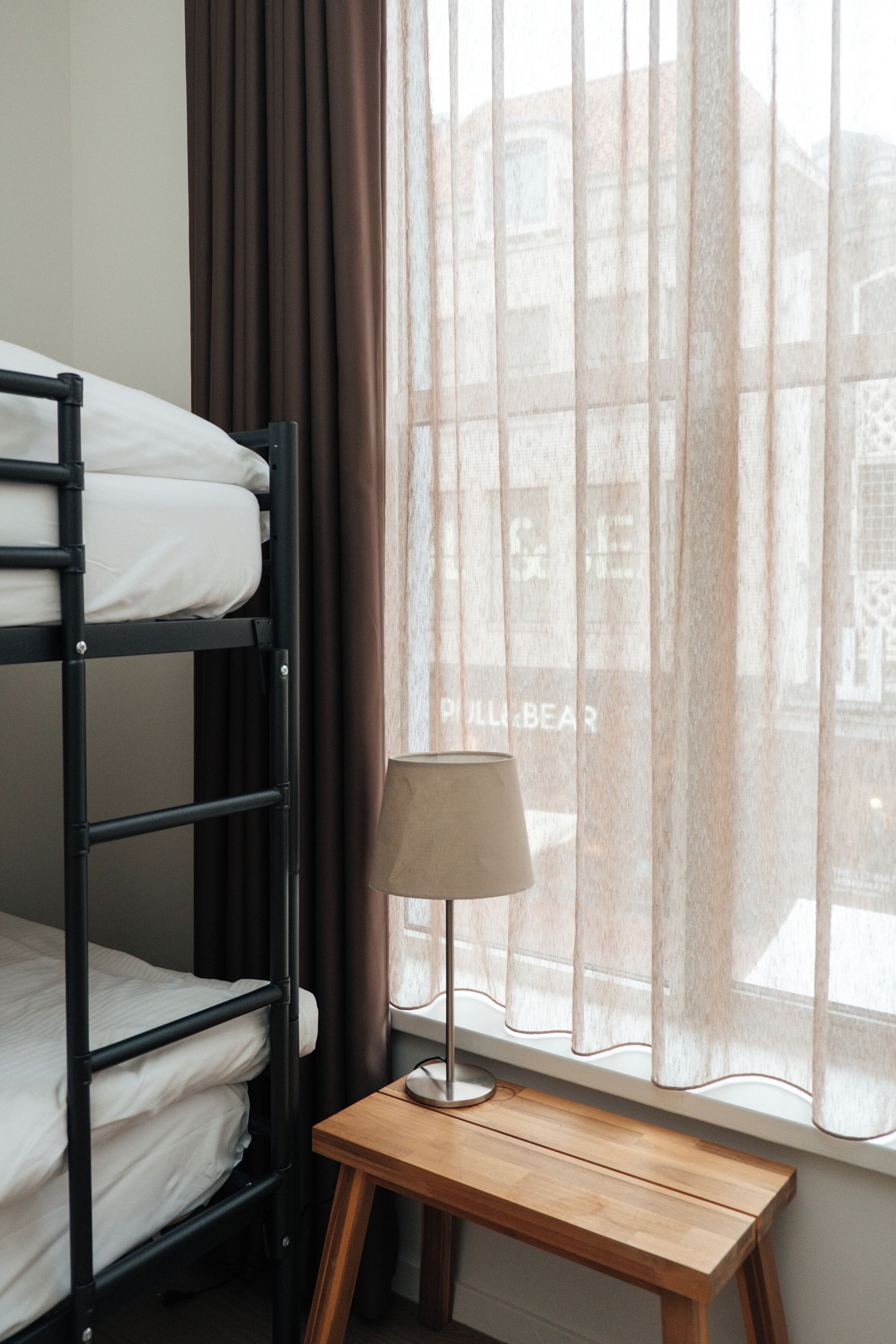 Un lit superposé, une table de chevet en bois, une lampe et une fenêtre avec des rideaux.