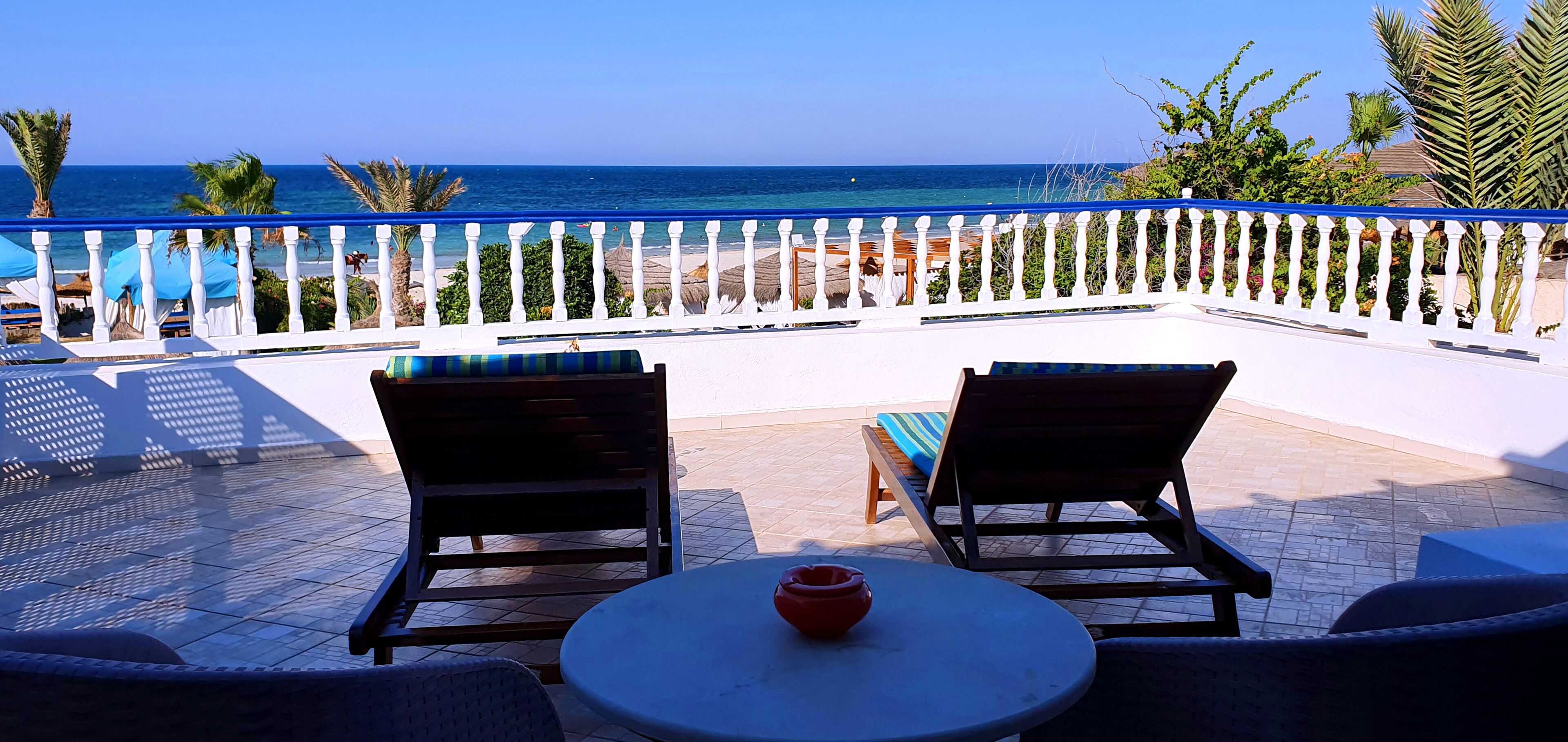 A balcony with two lounge chairs and a table, overlooking a beach with umbrellas and palm trees.