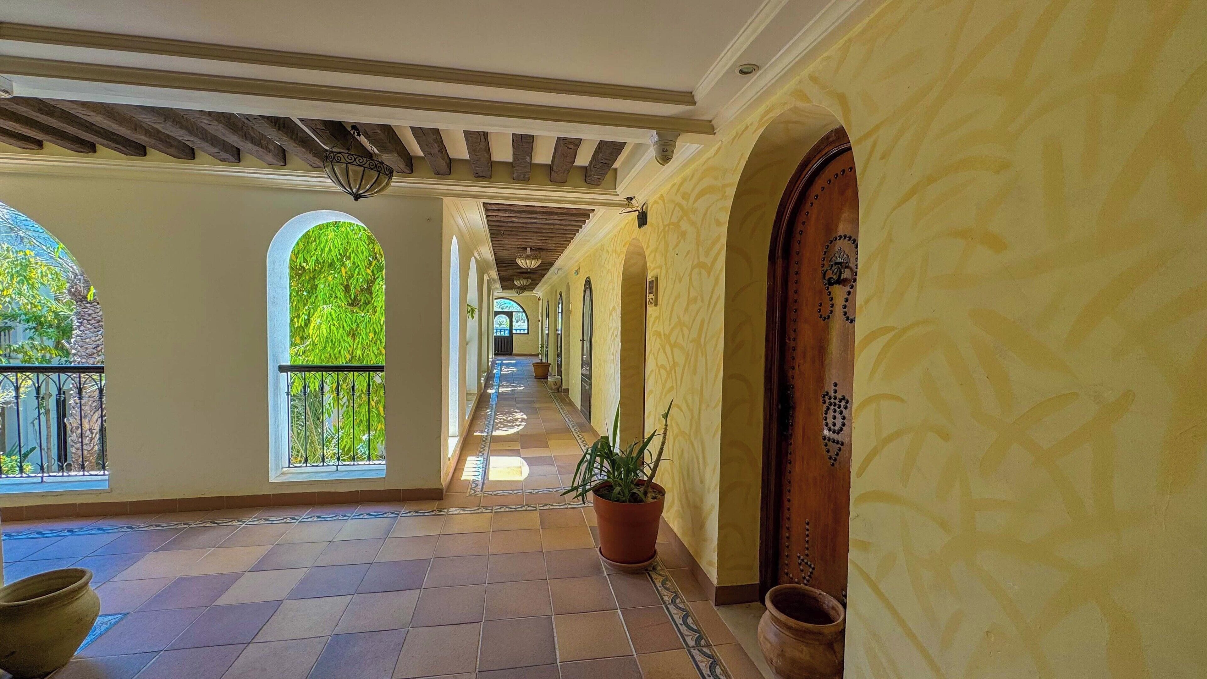 A corridor with tiled flooring, arched windows, and a potted plant.