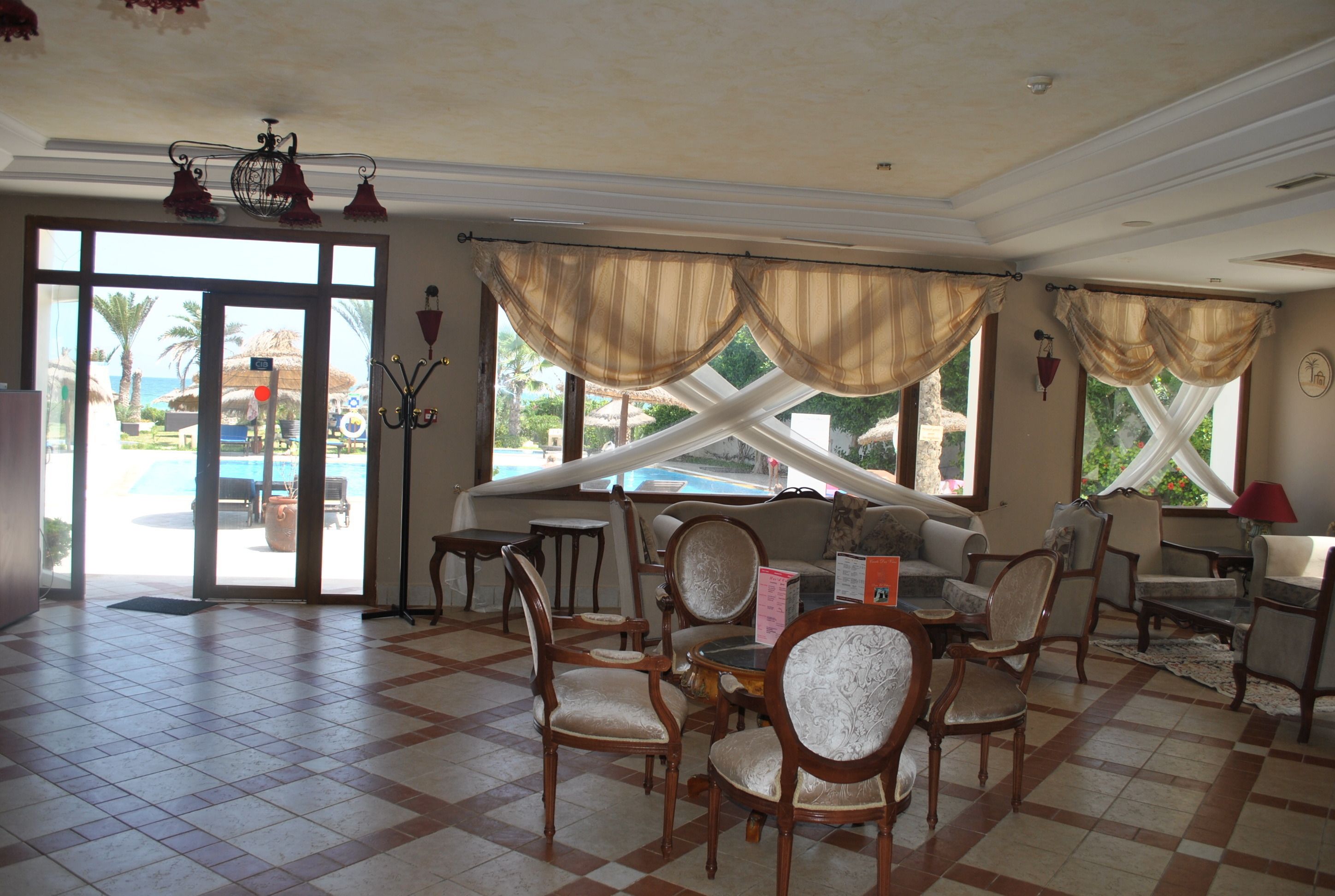 Eine großzügige Lobby mit Schachbrettmuster-Boden, Blick auf einen Pool durch große Fenster und klassischen Möbeln.