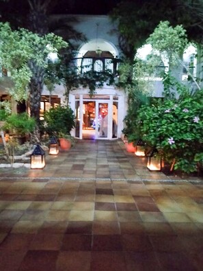 Un chemin menant à l’entrée d’un bâtiment, avec une porte en verre, le tout entouré de verdure et illuminé par des lanternes.