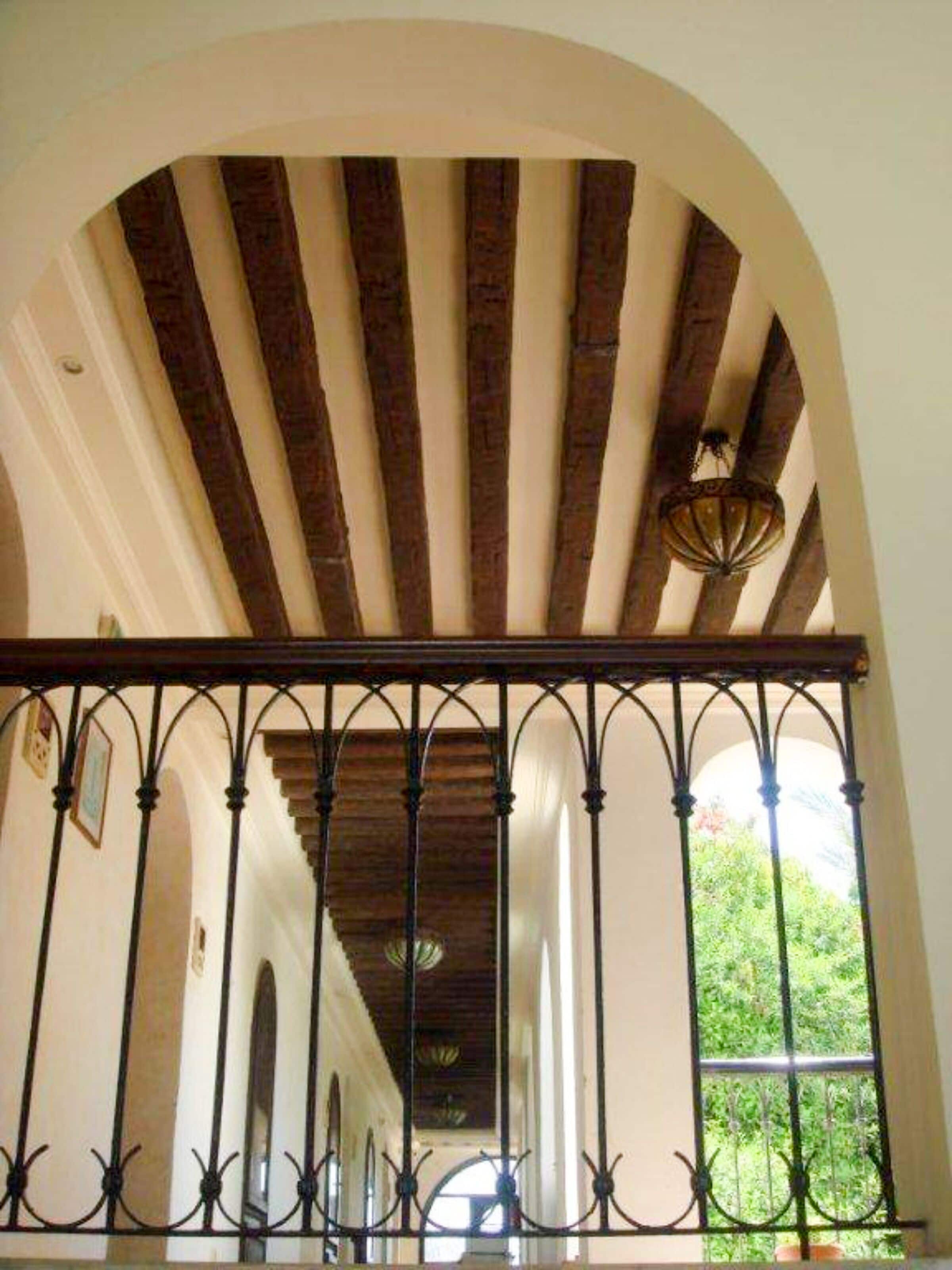 A corridor with a wooden ceiling, a wrought iron railing, and a view of greenery outside.