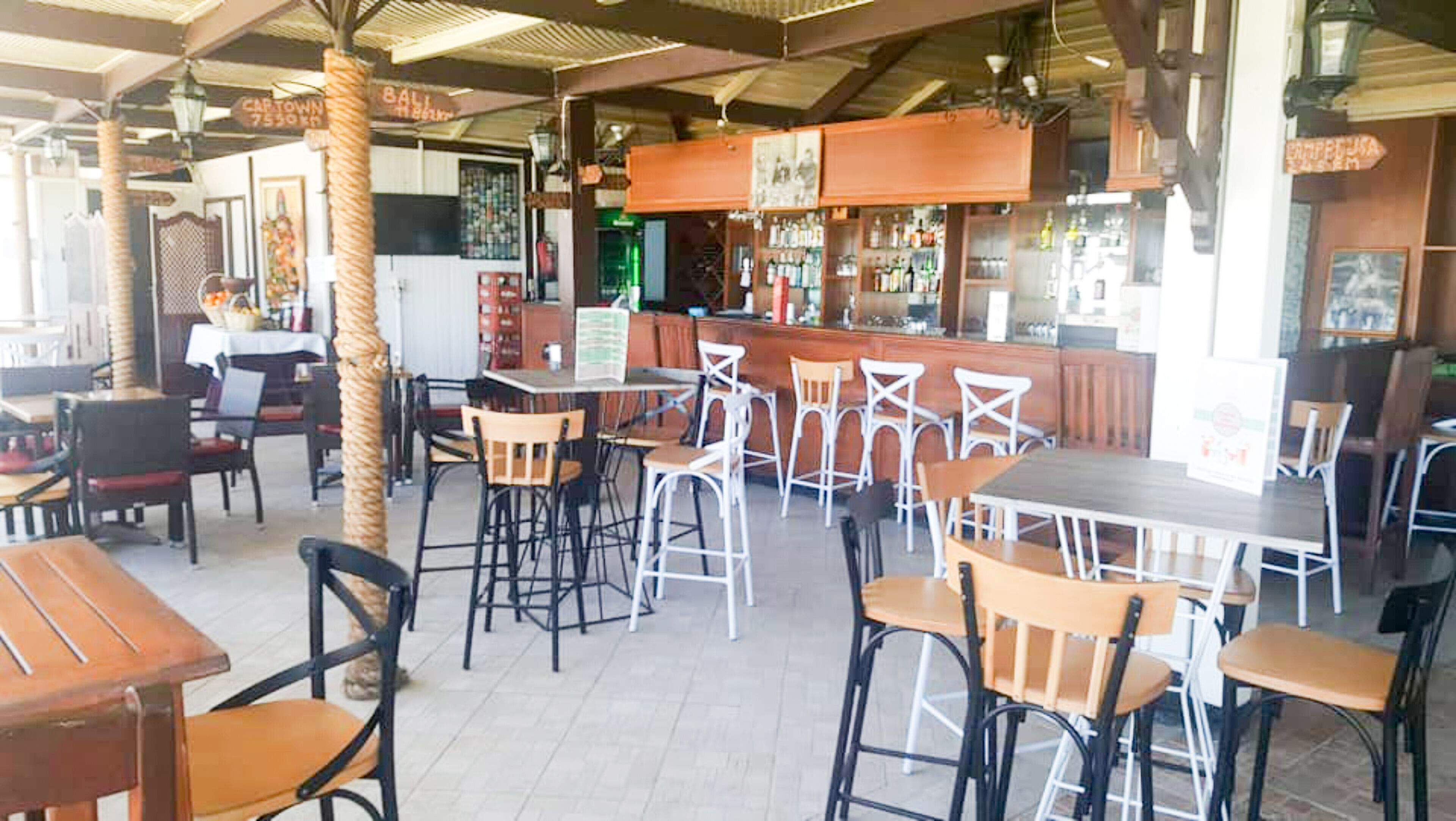 Un café al aire libre con mesas y sillas de madera, una zona de bar y una palmera tropical.